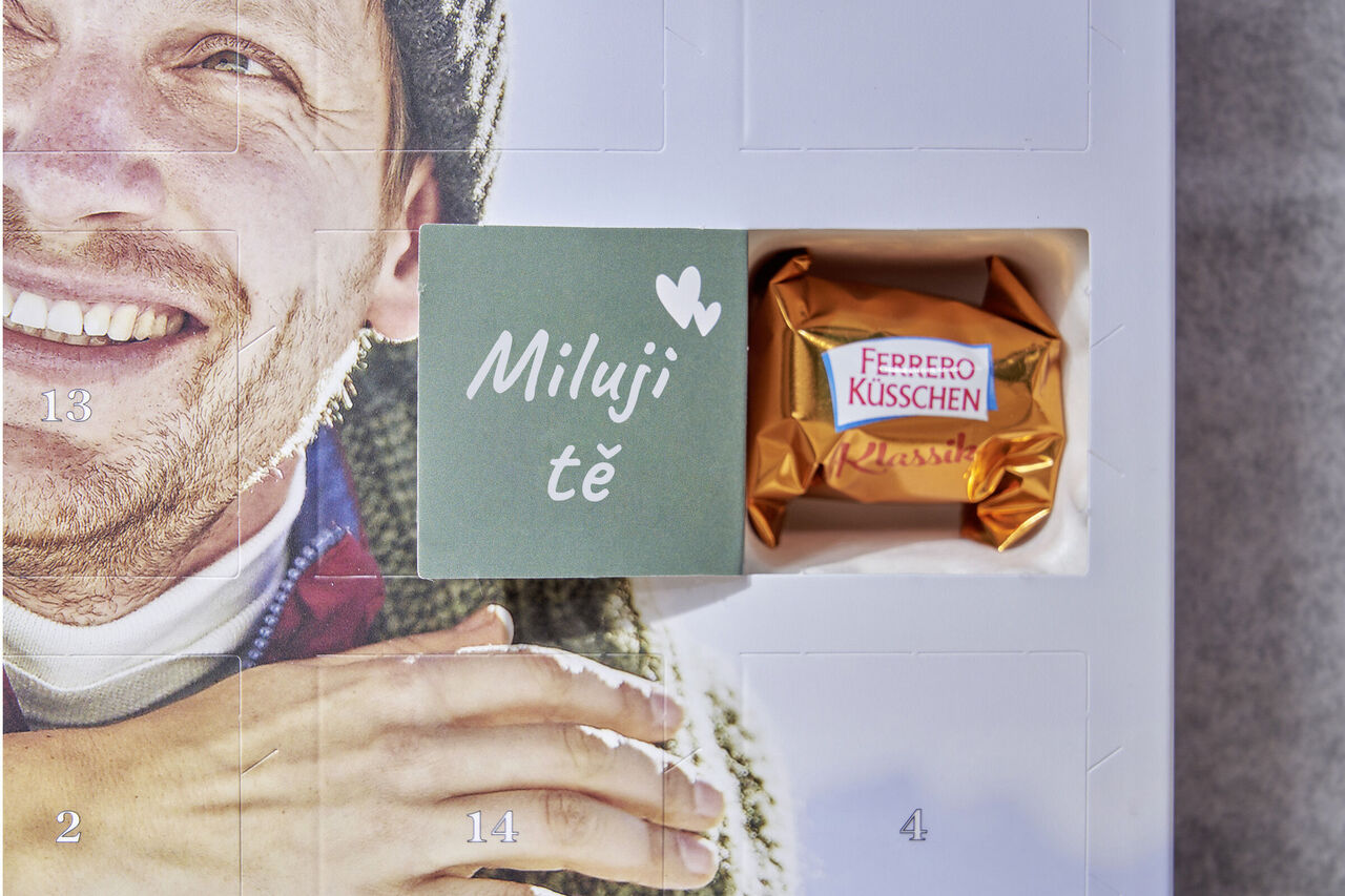 Na vnitřní straně otevřeného okénka adventního kalendáře je napsáno "Miluji tě". Vedle něj jsou dvě malá srdíčka. V okénku je Ferrero Küsschen.